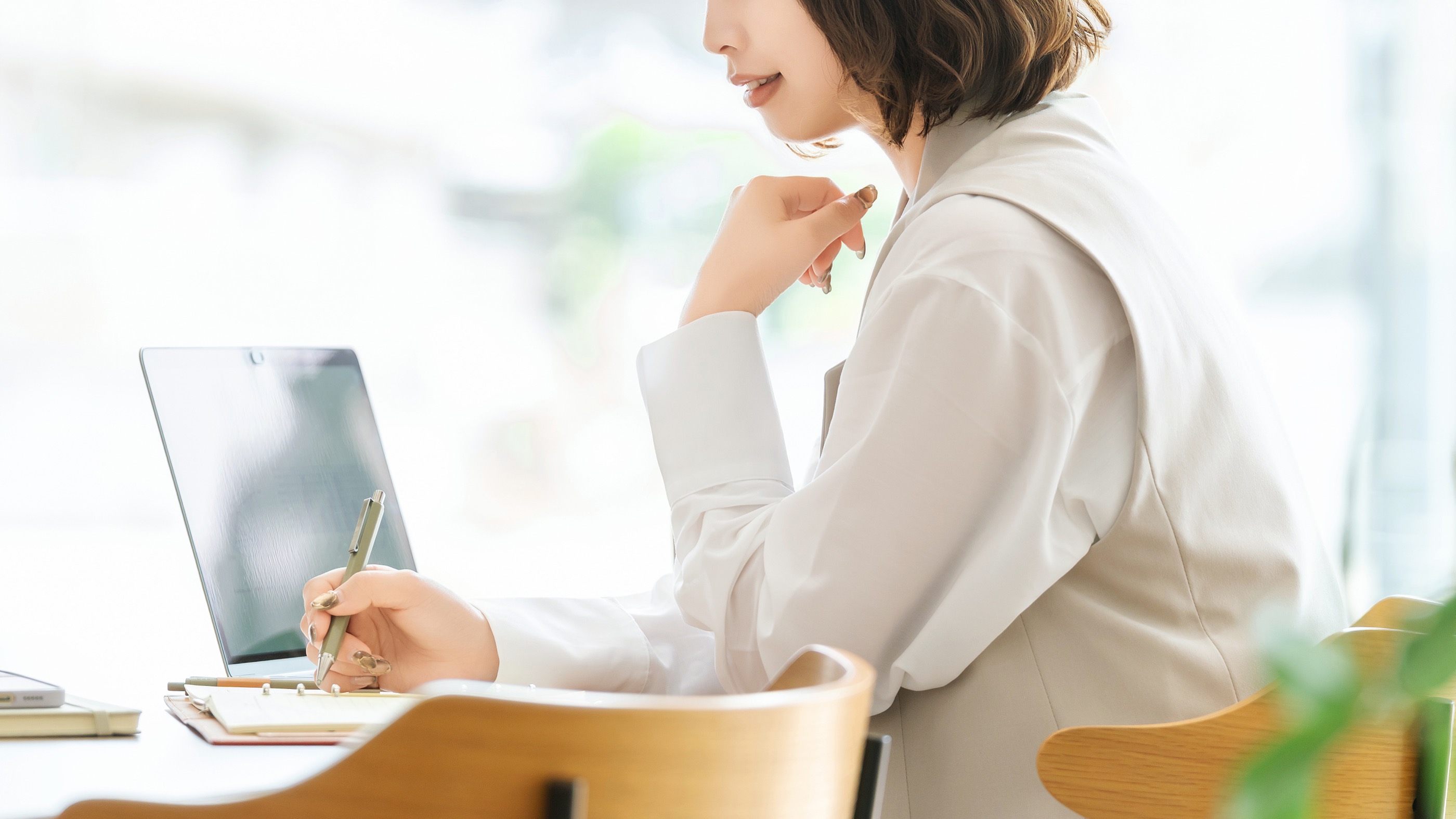 明るい窓辺でノートパソコンに向かい、考えごとをしている女性